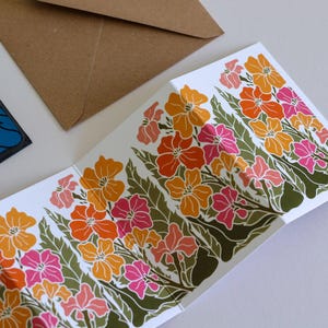 May include: A folded white greeting card featuring a repeating screen-printed pattern of orange, yellow, and pink flowers with dark green leaves. A brown paper envelope is positioned above the card, and a blue card corner is visible.