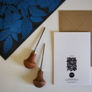 May include: A white card with a black and white illustration of a rose bush. The card has the text "Orig Rose" and "Adventures in Print" printed on it. The card is sitting on a brown envelope. Two wooden carving tools are on a white surface next to the card.