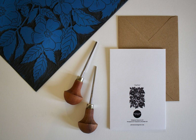 May include: A white card with a black and white illustration of a rose bush. The card has the text "Orig Rose" and "Adventures in Print" printed on it. The card is sitting on a brown envelope. Two wooden carving tools are on a white surface next to the card.