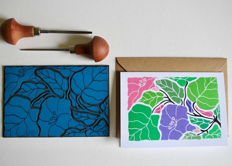 May include: A linoleum block print of a floral design with blue, green, pink, and purple flowers and leaves. The block print is on a white background. A card with the same design is printed in color on a white background.