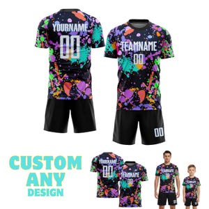 May include: Two matching soccer jerseys and shorts sets. The black jerseys have a colourful splatter design in green, purple, orange, and blue. The text "YOURNAME" and "TEAMNAME" are printed on the jerseys, along with the number "00". The shorts are black. The image also shows the text "CUSTOM ANY DESIGN".
