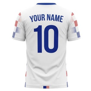 May include: White soccer jersey with blue accents and the text "YOUR NAME" above the number "10" in blue. The jersey has a blue collar and checkered patterns in red, white, and blue on the sleeves and hem.