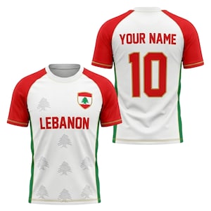 Könnte beinhalten: Weiß-rotes Fußballtrikot mit dem Wort "LEBANON" und einem Zedernbaum-Emblem auf der Vorderseite. Auf der Rückseite stehen die Nummer 10 und der Text "YOUR NAME" in Rot. Die Ärmel sind rot mit goldenen Zierleisten.