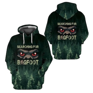 May include: A dark green hooded sweatshirt featuring a forest print, white drawstrings, and a graphic of glowing red eyes above the text "SEARCHING FOR BAGFOOT" with a small silhouette of a creature standing between the words.