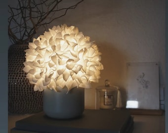 Könnte beinhalten: Eine weiße Keramik-Tischlampe mit einem einzigartigen, blumenförmigen Schirm. Die Lampe ist eingeschaltet und strahlt ein warmes Licht aus. Die Lampe steht auf einem schwarzen Buch mit dem Wort "SANI" auf dem Rücken.