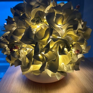 Könnte beinhalten: Eine weiße Papierblumenlampe mit weißem Sockel. Die Lampe ist eingeschaltet und das Licht scheint durch die Papierblumen.