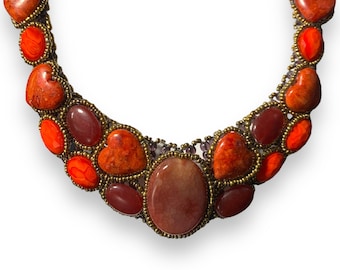 May include: A statement necklace with a large, oval-shaped, reddish-brown stone surrounded by smaller, heart-shaped, orange stones. The necklace is made with gold-toned metal and small, purple beads.