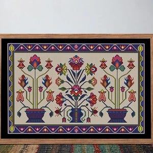 May include: Framed cross-stitch floral design featuring a central vase and two potted plants, all in vibrant reds, blues, and greens. The artwork is set against a beige background, framed in wood, and rests on a striped rug.