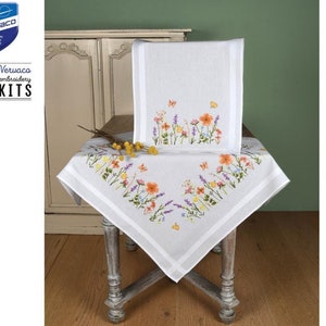 Puede incluir: Mantel y funda de silla bordados en blanco con un diseño floral con flores naranjas, moradas y amarillas, y mariposas. El mantel es cuadrado y tiene un borde blanco. También se muestra el texto "1 Stitch! Vervaco DIY Kits".