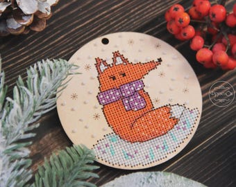 May include: A round wooden ornament featuring a cross-stitched fox wearing a purple scarf. The fox is orange with a white-tipped tail, set against a light beige background with snowflake accents. The ornament has a small hole for hanging.