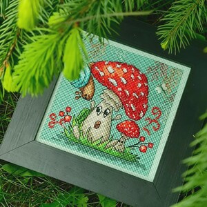 Op de afbeelding: Ingelijste kruissteekkunst van grillige paddenstoelen. De grote paddenstoel heeft een rode hoed met witte stippen. Het kunstwerk heeft een lichtgroene achtergrond en is omgeven door groen.