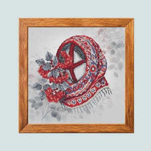 May include: A square cross-stitch embroidery featuring a red, white, and blue patterned headscarf draped over a cluster of red berries and grey leaves, all encased in a light brown wooden frame with a visible grain and a square profile.