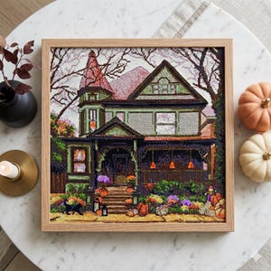 Puede incluir: Un bordado de punto de cruz enmarcado de una casa espeluznante con un exterior verde, un tejado rojo y un porche decorado para Halloween. La escena incluye calabazas, flores y un gato negro. La obra de arte está en un marco de madera clara.