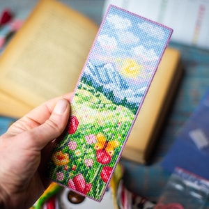 Könnte beinhalten: Handgefertigtes Lesezeichen mit Kreuzstichmuster. Das Lesezeichen zeigt eine Landschaft mit Bergen, Sonne und bunten Blumen. Der Rand ist lila. Im Hintergrund sind ein offenes Buch und Bastelmaterial zu sehen.