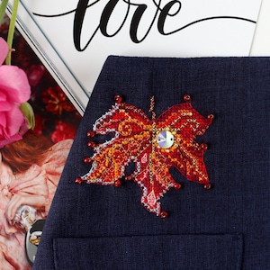 May include: A dark blue fabric with a red, orange, and yellow embroidered leaf with a sparkly centre. The leaf is sewn onto the fabric.