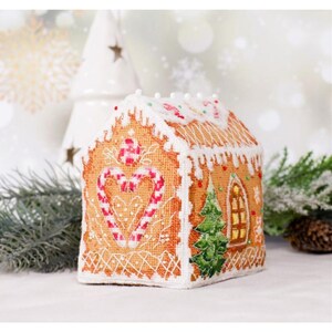 May include: A cross-stitched gingerbread house ornament featuring a heart-shaped candy cane design on the front, a green pine tree, and a yellow window on the side. The roof is decorated with white icing details and small red and green beads.