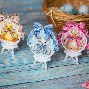 May include: Three embroidered Easter egg ornaments on small white easels. The ornaments feature a duckling, a bunny, and a chick, each with a bow. A basket with Easter eggs and embroidery threads are in the background.