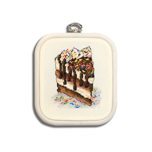 Può includere: Un ricamo a punto croce di una fetta di torta al cioccolato con glassa bianca e codette colorate. La torta è incorniciata in una cornice di legno bianca.