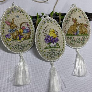 Op de afbeelding: Drie paasei-vormige ornamenten met kruissteekontwerpen. Elk heeft een andere lentescène: een konijn met een mand met eieren, een kuiken in een bloempot en twee konijnen. Ze hebben witte kwasten en zijn afgewerkt met wit koord.