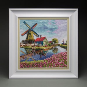 May include: A framed cross-stitch artwork depicting a colourful windmill with a green roof and white blades, a small green house with a red roof, and a canal with pink flowers in the foreground.