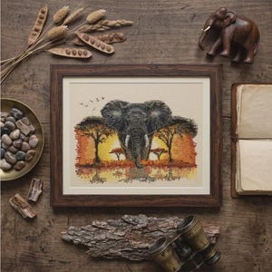 May include: A framed cross-stitch embroidery featuring a detailed elephant head centered against a vibrant orange and yellow sunset landscape with silhouetted acacia trees and a small flock of birds flying in the upper left corner of the scene.