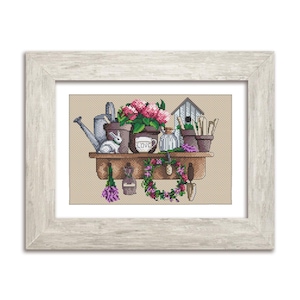 May include: Cross-stitch artwork in a light wood frame. The image shows a garden scene with a shelf holding potted flowers, a watering can, a bunny, and gardening tools. A wreath and lavender hang below. The word "LOVE" is on a mug.