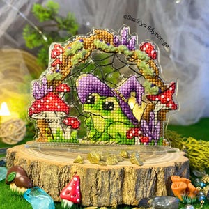 Op de afbeelding: Een kruissteekontwerp van een kikker met een paarse heksenhoed, omringd door rode en witte paddenstoelen en paarse kristallen. Het ontwerp is geplaatst in een helder acryl frame, op een houten basis. De tekst "Suzir'ya Edynoroga" is zichtbaar.