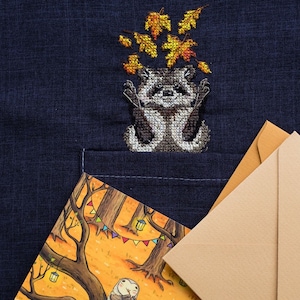 May include: A cross-stitch design of a raccoon holding a bunch of yellow and orange leaves. The design is on a dark blue fabric. A brown envelope with a wax seal is in the background.