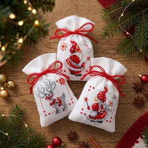 Peut inclure: Trois petits sacs en tissu blanc avec des cordons de serrage rouges, ornés de broderies au point de croix représentant un père Noël, un renne et un gnome. Chaque sachet est décoré de petits motifs d'étoiles rouges sur le devant.