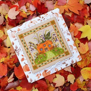May include: A cross-stitch design depicting an orange pumpkin house with a green lawn and a picket fence. The design is framed with a decorative border and set on white fabric with a pumpkin pattern. The background is filled with colourful autumn leaves.