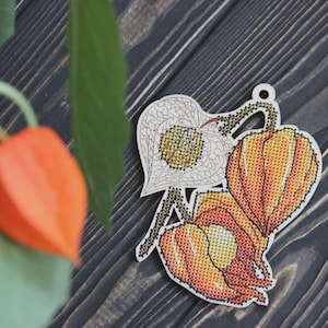 Könnte beinhalten: Holzornament mit Kreuzstichdesign von orangefarbenen und gelben chinesischen Lampionblumen. Das Ornament hat ein kleines Loch zum Aufhängen. Der Hintergrund ist eine dunkle Holzoberfläche.