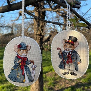 Puede incluir: Dos adornos ovalados bordados colgando de una rama. Uno presenta un ratón con vestido y sombrero azules, bufanda roja y paraguas. El otro representa un ratón con abrigo, chaleco y sombrero de copa, sosteniendo un bastón.