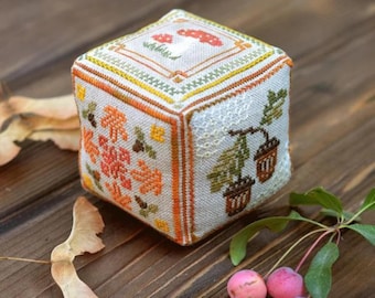 May include: A white fabric cube with cross-stitch embroidery depicting autumnal scenes. The cube features acorns, mushrooms, and leaves in shades of orange, brown, and green.