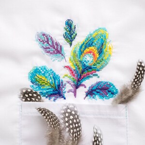 Könnte beinhalten: Weißer Stoff mit einem bunten gestickten Muster aus Federn. Ein paar echte Federn mit schwarzen und weißen Punkten befinden sich in der rechten unteren Ecke des Bildes.