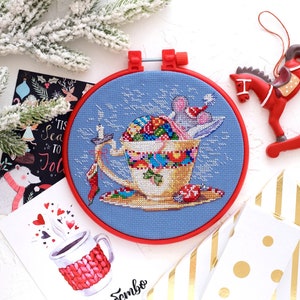 May include: A blue and white cross-stitch embroidery of a mouse in a teacup with a red Santa hat. The teacup is filled with colorful ornaments and candy. The embroidery is framed in a red hoop.