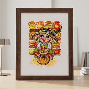 May include: A framed cross-stitch embroidery depicting a gnome wearing a green hat and apron, sitting at a pottery wheel. The gnome is surrounded by wooden shelves filled with various colorful ceramic bowls and pitchers on a white fabric background.