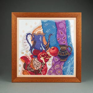 May include: A framed cross-stitch embroidery depicting a still life with a blue teapot, a small dark pitcher, and two pomegranates. The composition includes draped blue and purple patterned fabrics against a light background with subtle dots.