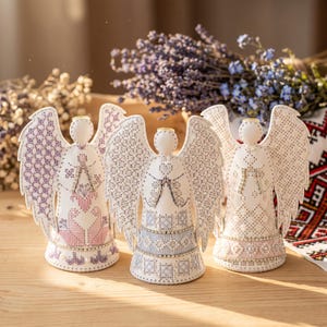 May include: Three white angel figurines featuring intricate embroidery and beadwork. Each angel has detailed wings and a dress decorated with patterns in shades of pink, blue, and silver. The figures are standing upright with a circular base.