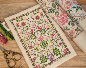May include: A completed cross-stitch project featuring a floral and bird design in shades of pink, green, and purple. Two rolled cross-stitch patterns are visible in the background. Embroidery floss and scissors are also present, suggesting a crafting theme.