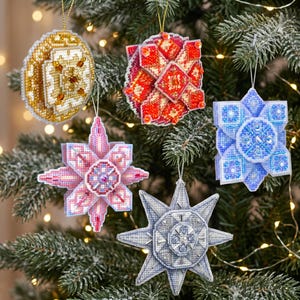 May include: Five three-dimensional geometric Christmas ornaments crafted from plastic canvas and beads. The collection includes a gold circular piece, a red and white square, a pink star, a blue square, and a grey star, all hanging by gold strings.