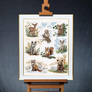 May include: A framed cross-stitch embroidery featuring a variety of woodland animals, including a deer fawn, owl, wolf pup, squirrel, rabbit, and bear cub. The animals are depicted in a whimsical style against a white background with blue clouds.