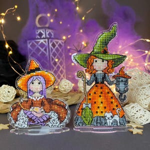 May include: Two cross-stitched witch figurines. One witch wears an orange and purple dress and hat. The other witch wears an orange and black dress and green hat, holding a lantern and broom. A white cat is at her feet. The background has purple accents and fairy lights.