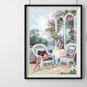 May include: Cross-stitch artwork framed in black depicts a serene outdoor scene. Two white wicker chairs with cushions sit on a stone patio, facing a table with a pink tablecloth. A pink parasol and flowers add color to the scene.