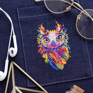 Puede incluir: Un diseño de búho bordado de colores sobre un bolsillo de tela vaquera azul oscuro. El búho está cosido con varios colores, incluyendo rosa, amarillo, naranja, verde y azul. Hay gafas con montura dorada, un soporte geométrico dorado y un pequeño frasco de vidrio lleno de purpurina dorada sobre la tela.
