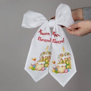 Könnte beinhalten: Weißes Oster-Leinentuch mit gestickten Kuchen, Kerzen und bunten Eiern. Das Tuch hat roten Text und ist zu einer Schleife gebunden. Eine festliche Dekoration für die Osterfeiertage.