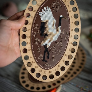 Può includere: Un organizzatore di fili in legno con 32 fori per riporre il filo da ricamo. L'organizzatore ha un pannello centrale in pelle marrone con un design di gru ricamato in bianco e marrone. I numeri da 16 a 32 sono visibili sull'organizzatore.