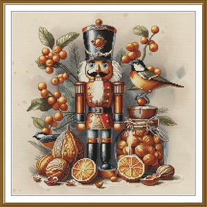 Op de afbeelding: Een kruissteekpatroon met een centrale notenkrakerfiguur in een zwarte hoed en rood uniform, omringd door oranje bessen, dennennaalden, een kleine vogel, een glazen pot gevuld met walnoten en schijfjes sinaasappel op een beige achtergrond.