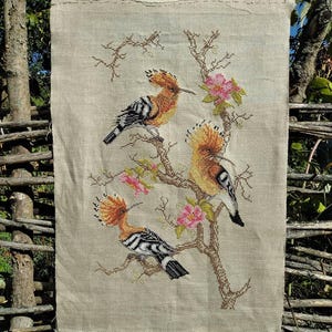 May include: A cross-stitch tapestry depicting three hoopoe birds on a flowering branch. The birds have orange crests, black and white striped wings, set against a neutral linen backdrop. Pink flowers and brown branches add colour to the design.