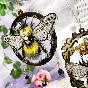 May include: A decorative bee ornament with a gold-colored frame. The bee has yellow and black fur, clear wings, and a jeweled body. Black teardrop-shaped beads hang from the frame. The ornament is likely for home decor or a holiday.