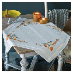 Könnte beinhalten: Eine weiße Tischdecke mit einem gestickten Muster aus Vögeln und Blättern in Orange-, Gelb- und Blautönen. Zwei Kerzen in braunen Haltern stehen auf dem Tisch.